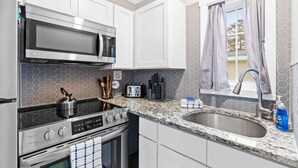 A modern kitchen with stainless steel appliances, a granite countertop, and a window with curtains. - Cozy Cottage Walking Distance to the Beach With Coffee Bar (Oak Island)
