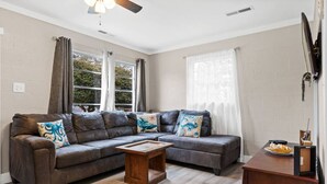 A living room with a brown leather sofa, two matching armchairs, a wooden coffee table, and a TV on a wooden stand. - Cozy Cottage Walking Distance to the Beach With Coffee Bar (Oak Island)