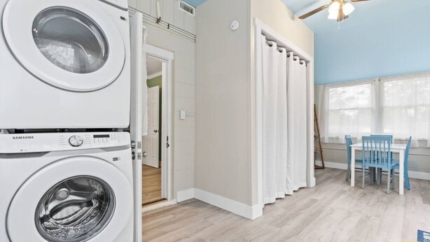 A laundry room with stacked washers and dryers, a dining area with blue chairs, and a ceiling fan. - Cozy Cottage Walking Distance to the Beach With Coffee Bar (Oak Island)