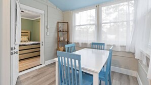 A room with a white table and blue chairs, a wooden bed, and a window with sheer curtains. - Cozy Cottage Walking Distance to the Beach With Coffee Bar (Oak Island)