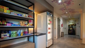 Un pasillo moderno de hotel con un refrigerador lleno de bebidas, estantes con artículos de aseo y un mural en la pared.