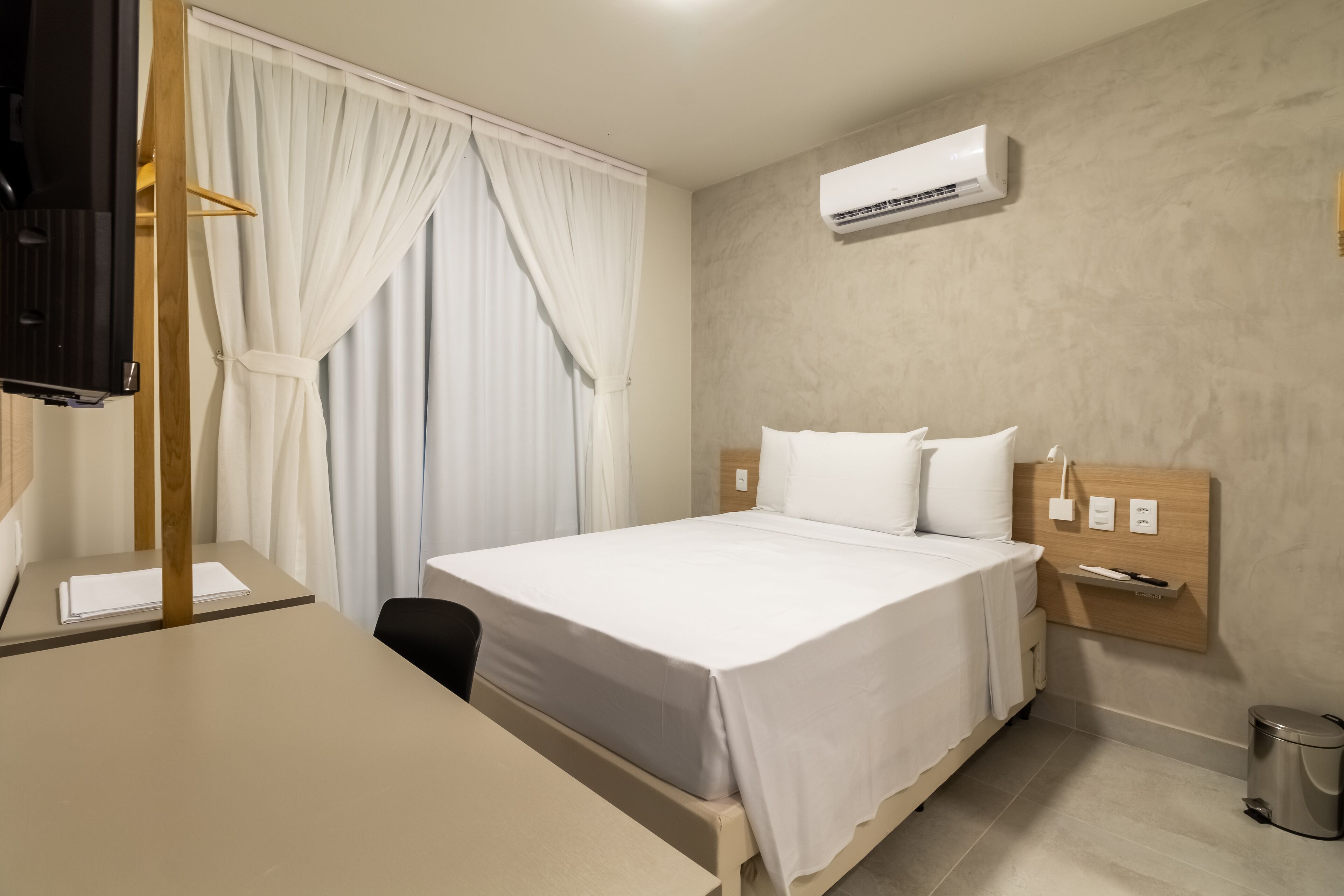 A hotel room with a bed, a desk, a chair, an air conditioning unit, and a trash bin.