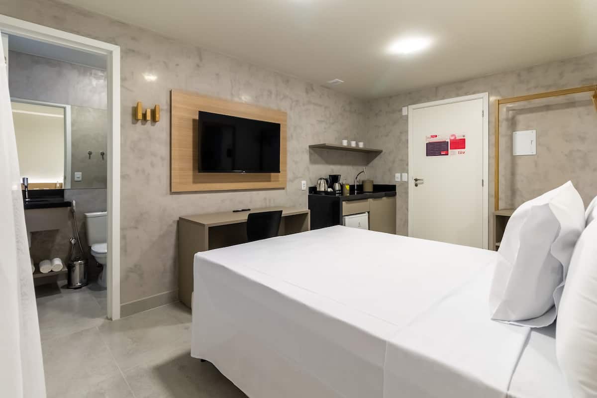 A hotel room with a bed, a desk, a TV mounted on the wall, a small kitchenette, and a bathroom visible through an open door.