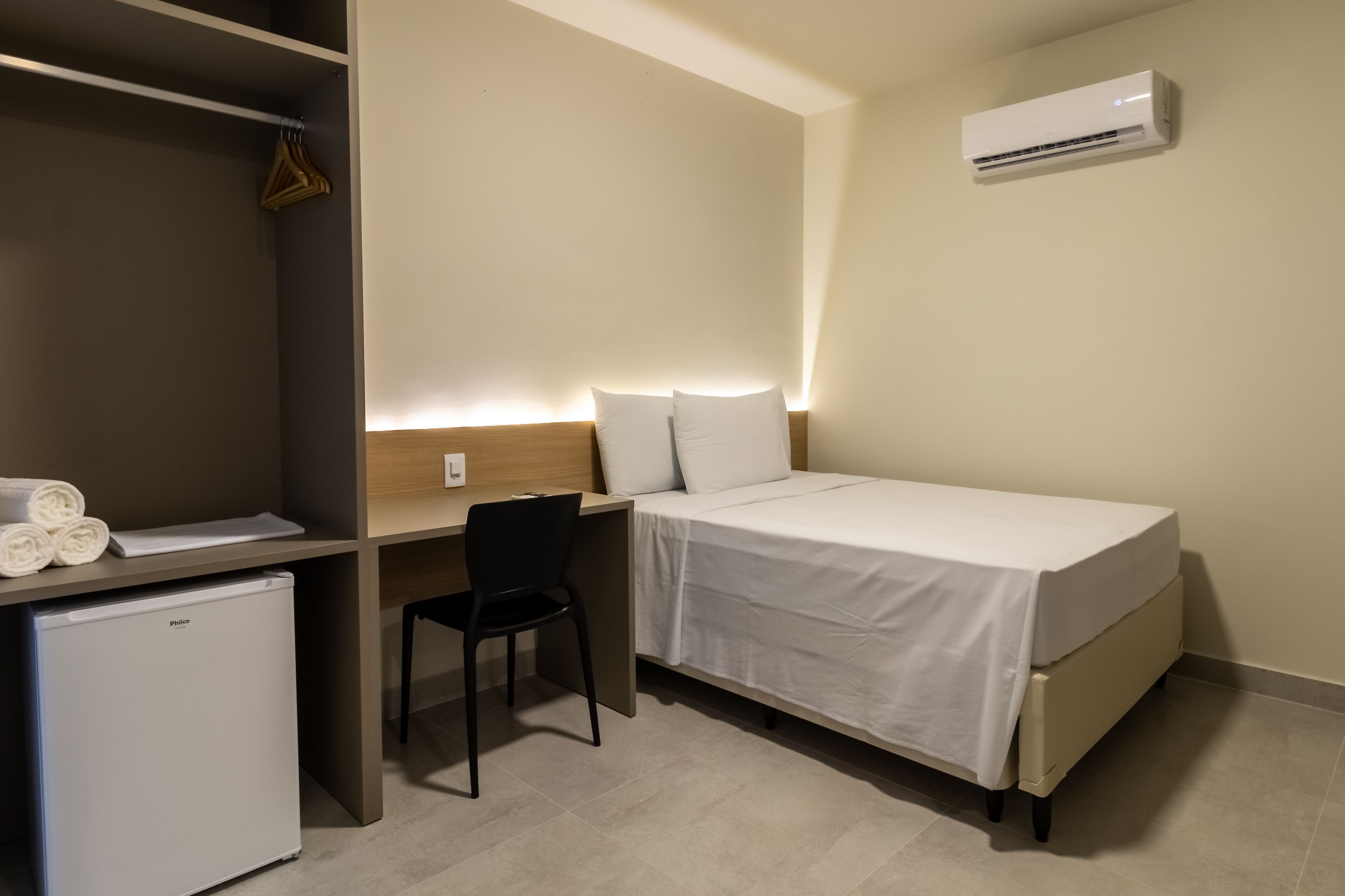 Habitación de hotel compacta con una cama individual, un escritorio con silla, un refrigerador pequeño y un aire acondicionado instalado en la pared.