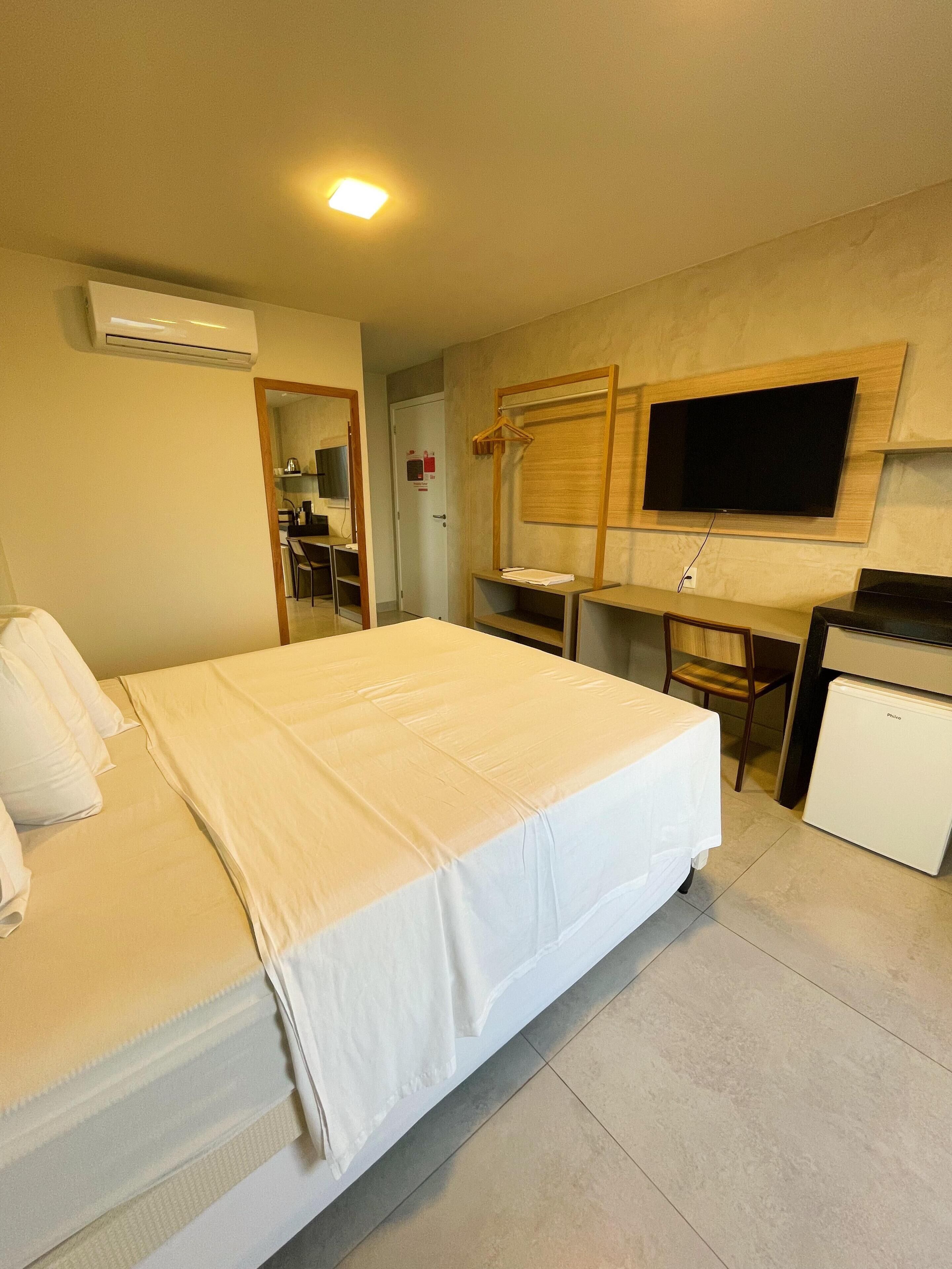 Quarto de hotel com cama, escrivaninha, televisão e mini-geladeira.