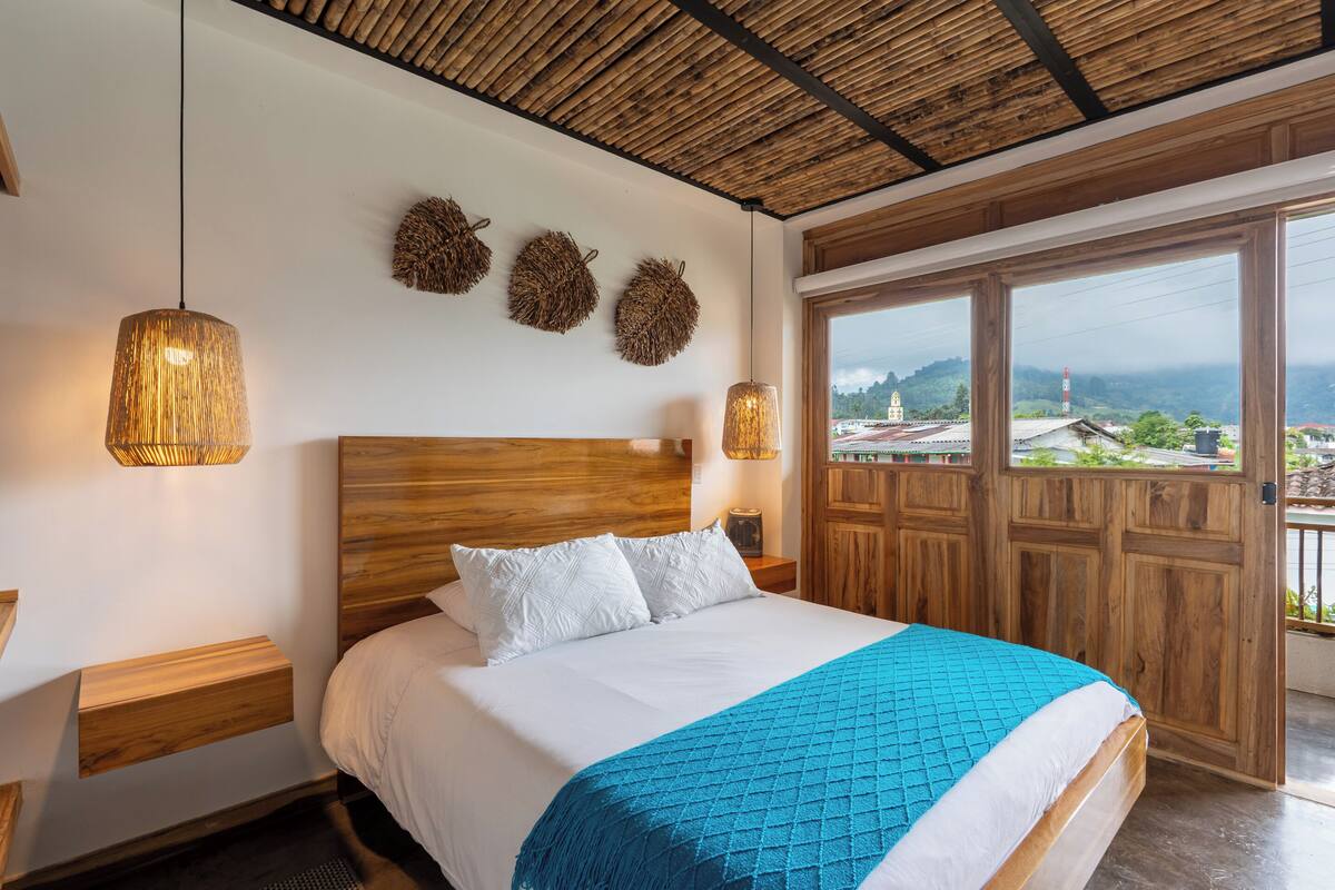 A bedroom with a wooden headboard, a large bed with a blue blanket, two bedside lamps, and a view of a town through a wooden door.
