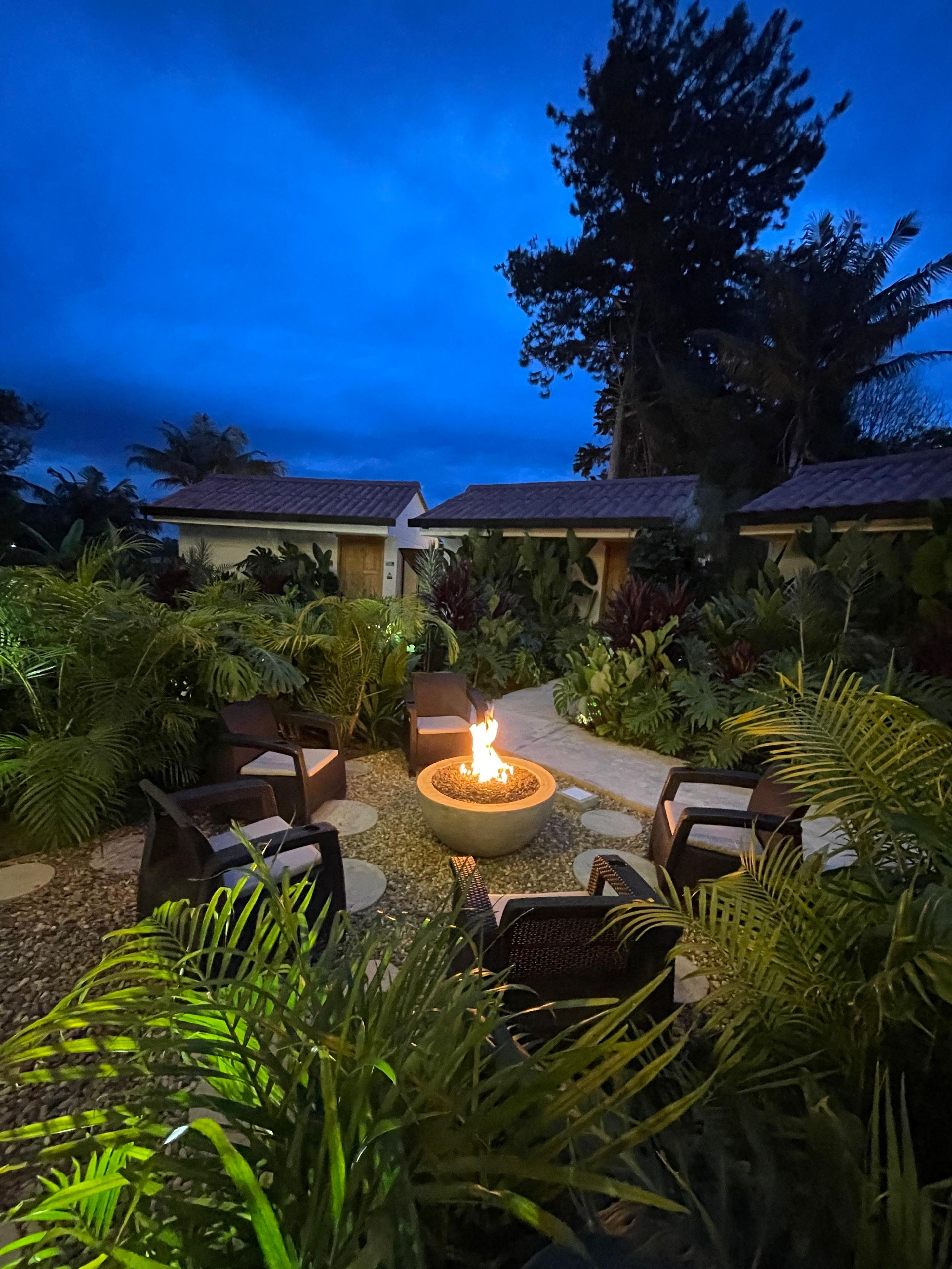 A patio area with a fire pit surrounded by outdoor furniture, lush greenery, and a pathway leading to a building with a tiled roof.