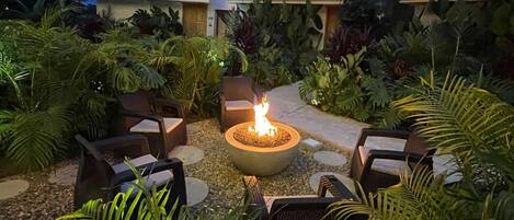 A patio area with a fire pit surrounded by outdoor furniture, lush greenery, and a pathway leading to a building with a tiled roof.