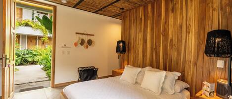 A bedroom with a wooden ceiling, a bed with white bedding, a bedside table with a lamp, and a view of a garden through an open door.