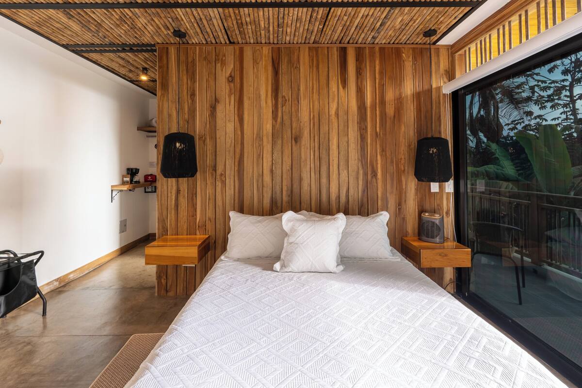 A bedroom with a wooden panel wall, a large bed with white bedding, wooden nightstands, and a window with a view of greenery.