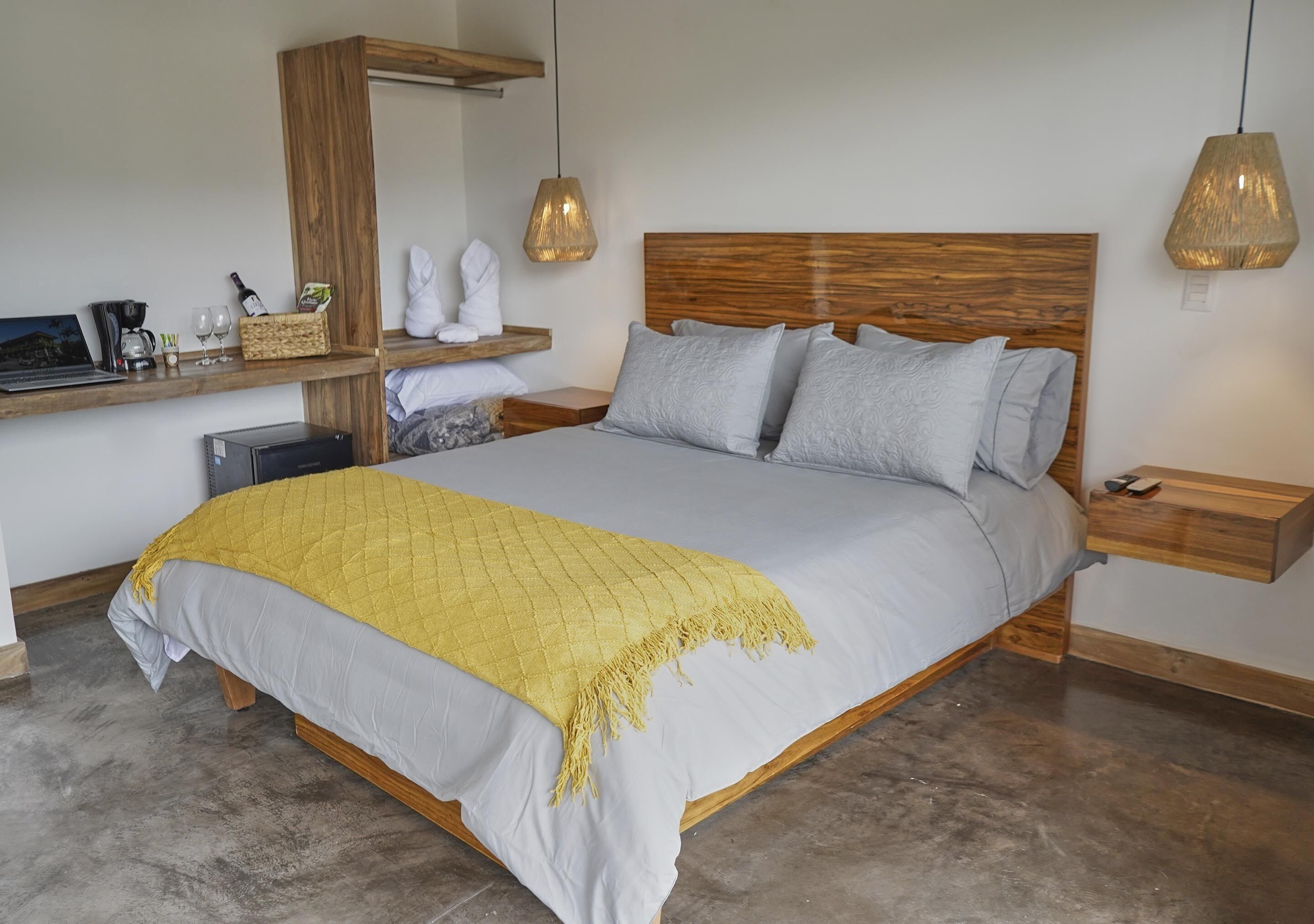Een slaapkamer met een houten hoofdbord, een bed met een gele deken, een nachtkastje met een lamp en een plank met een laptop en decoratieve objecten.