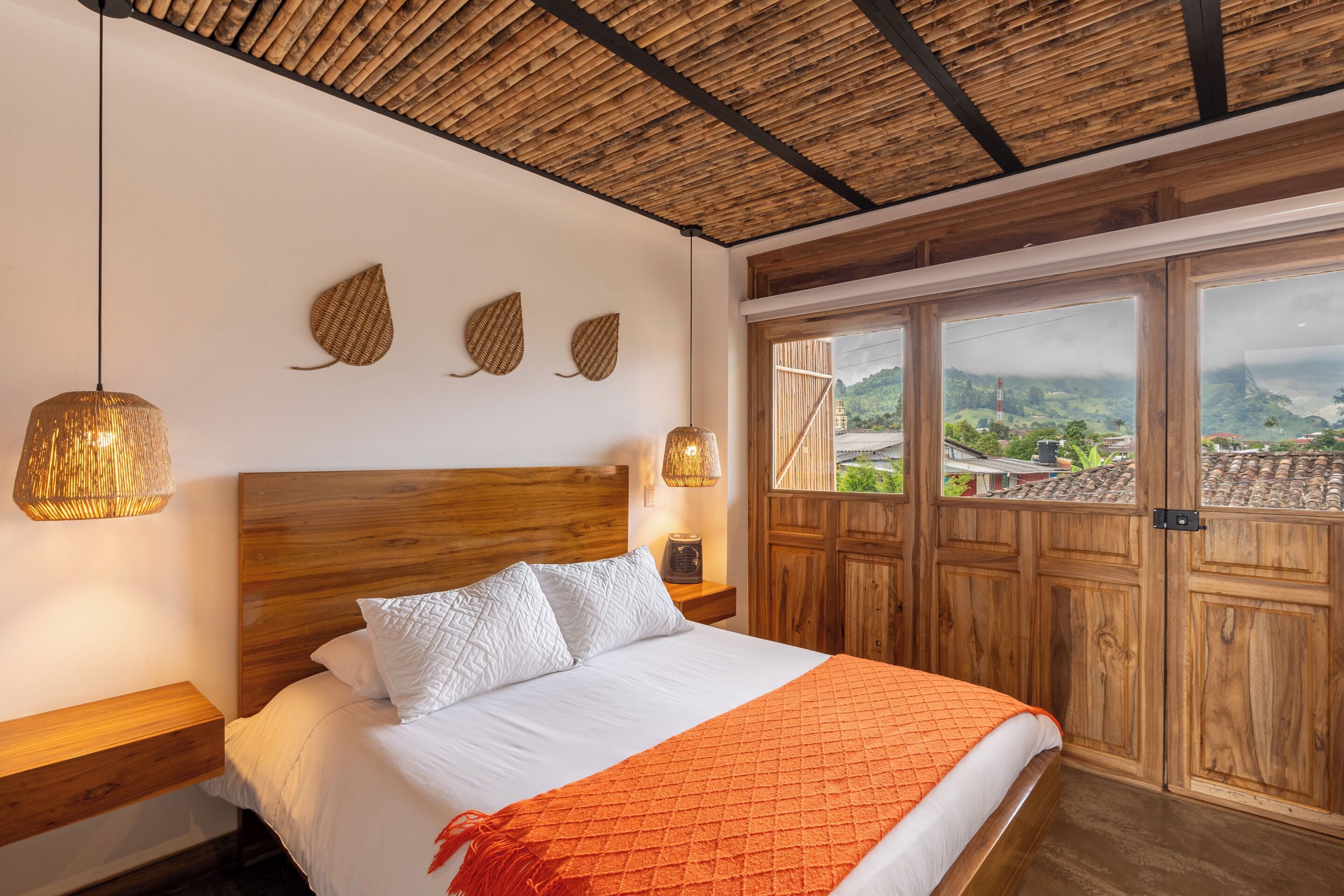 Une chambre à coucher avec une tête de lit en bois, un lit recouvert de draps blancs, un plaid orange et une vue sur un paysage montagneux, visible à travers de grandes fenêtres.