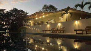 Ein Poolbereich mit Liegestühlen und einer überdachten Sitzgelegenheit, umgeben von Bäumen und einem klaren Pool, der die Lichter reflektiert.