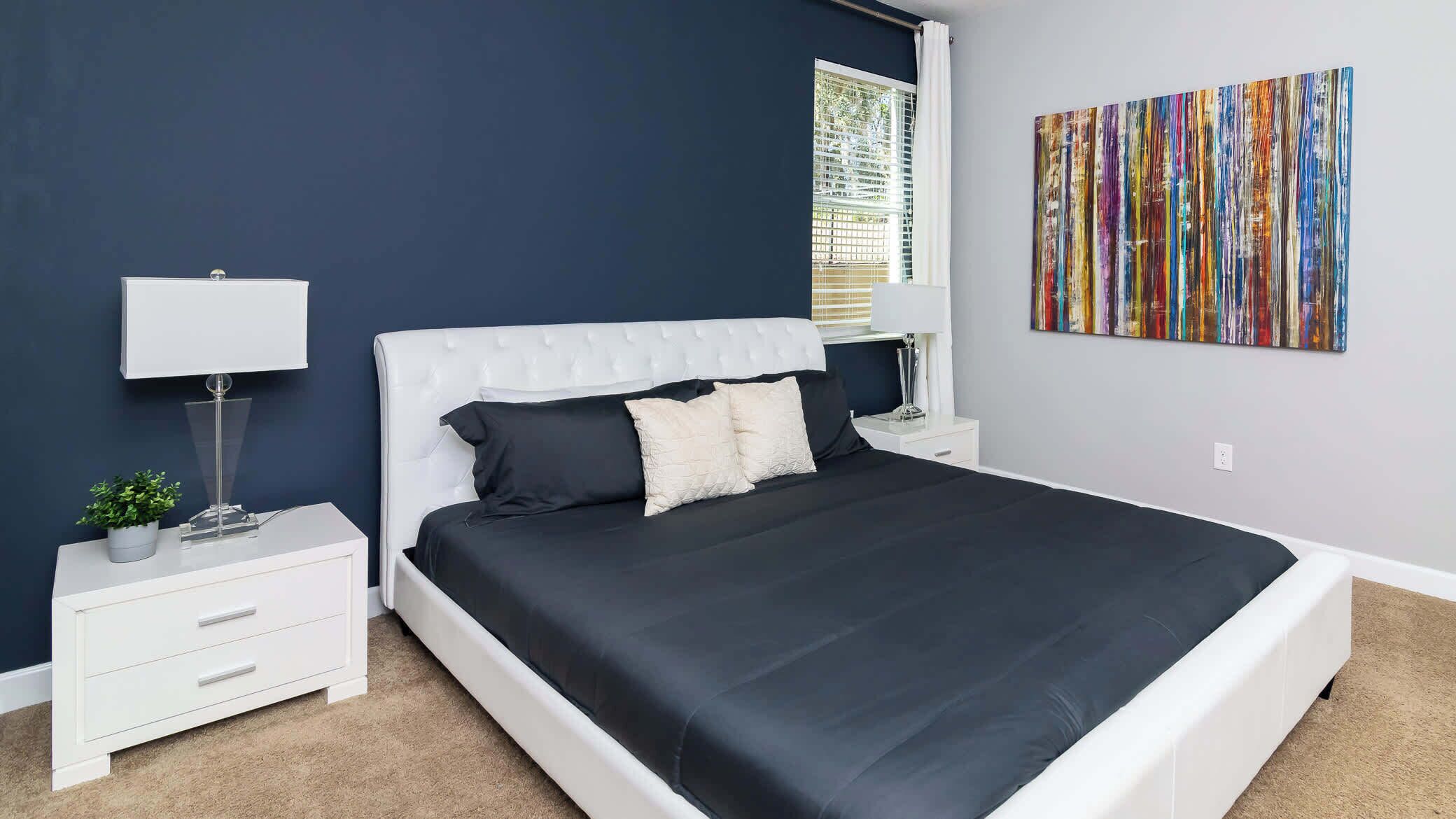 A modern bedroom with a large bed, a nightstand, a lamp, a potted plant, and a colorful abstract painting on the wall.