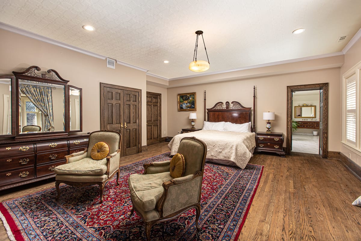 A spacious bedroom with a large bed, two armchairs, a dresser, and a mirror.