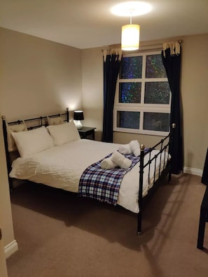 Una cama bien hecha con una manta escocesa y toallas enrolladas, una lámpara de noche y una ventana con cortinas.