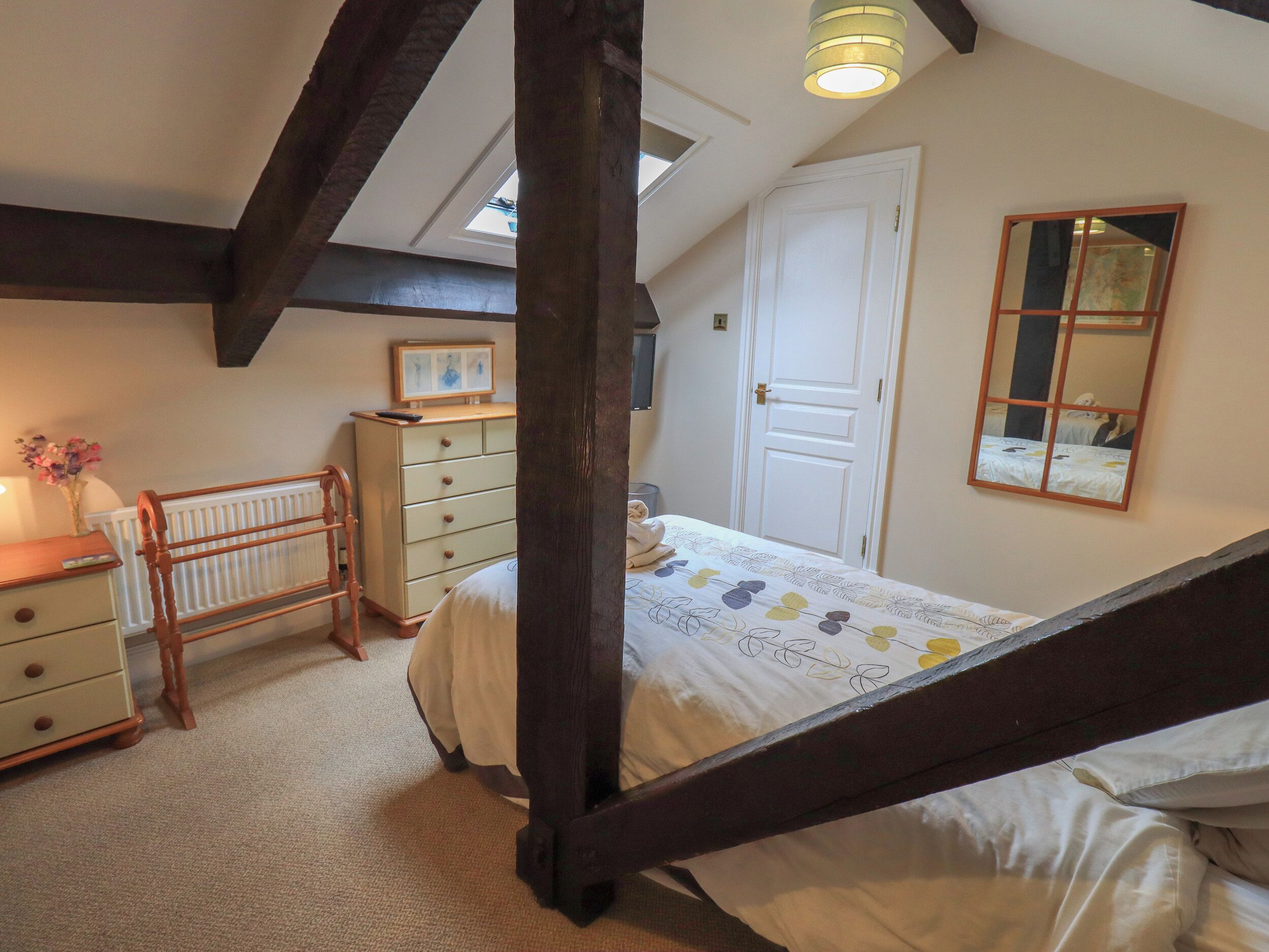 A bedroom with a sloped ceiling, a bed with a floral bedspread, a dresser, a mirror, and a window with a view of the sky.