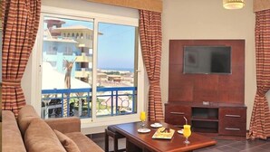 A living room with a sofa, a dining table with breakfast items, a TV, and a balcony with a view of buildings and the sea. - Porto Sharm Hotel Apartments (Sharm El Sheikh)