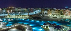 A large, illuminated swimming pool complex with multiple sections and a central island, surrounded by hotel buildings with multiple floors and balconies.