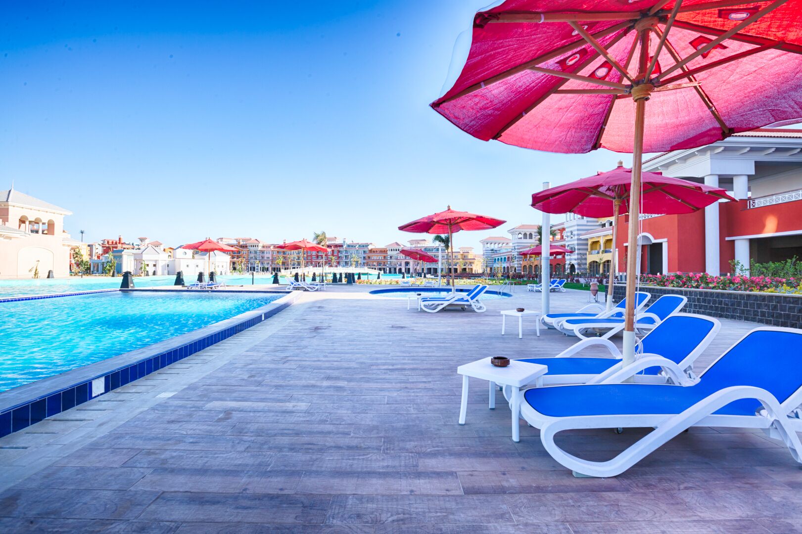 A poolside area with blue lounge chairs and red umbrellas, surrounded by buildings with balconies.
