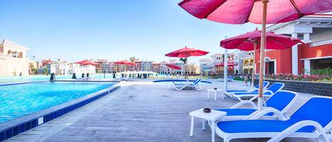 A poolside area with blue lounge chairs and red umbrellas, surrounded by buildings with balconies.