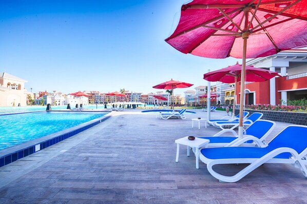 A poolside area with blue lounge chairs and red umbrellas, surrounded by buildings with balconies.