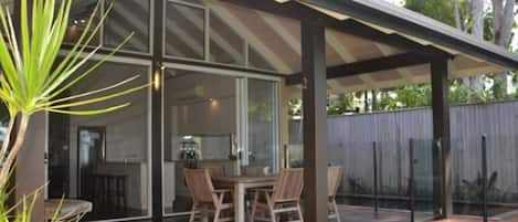 Ein überdachter Außenbereich mit Tisch und Stühlen, eine Glastür, die zu einem Innenraum führt, und ein Holzzaun im Hintergrund.