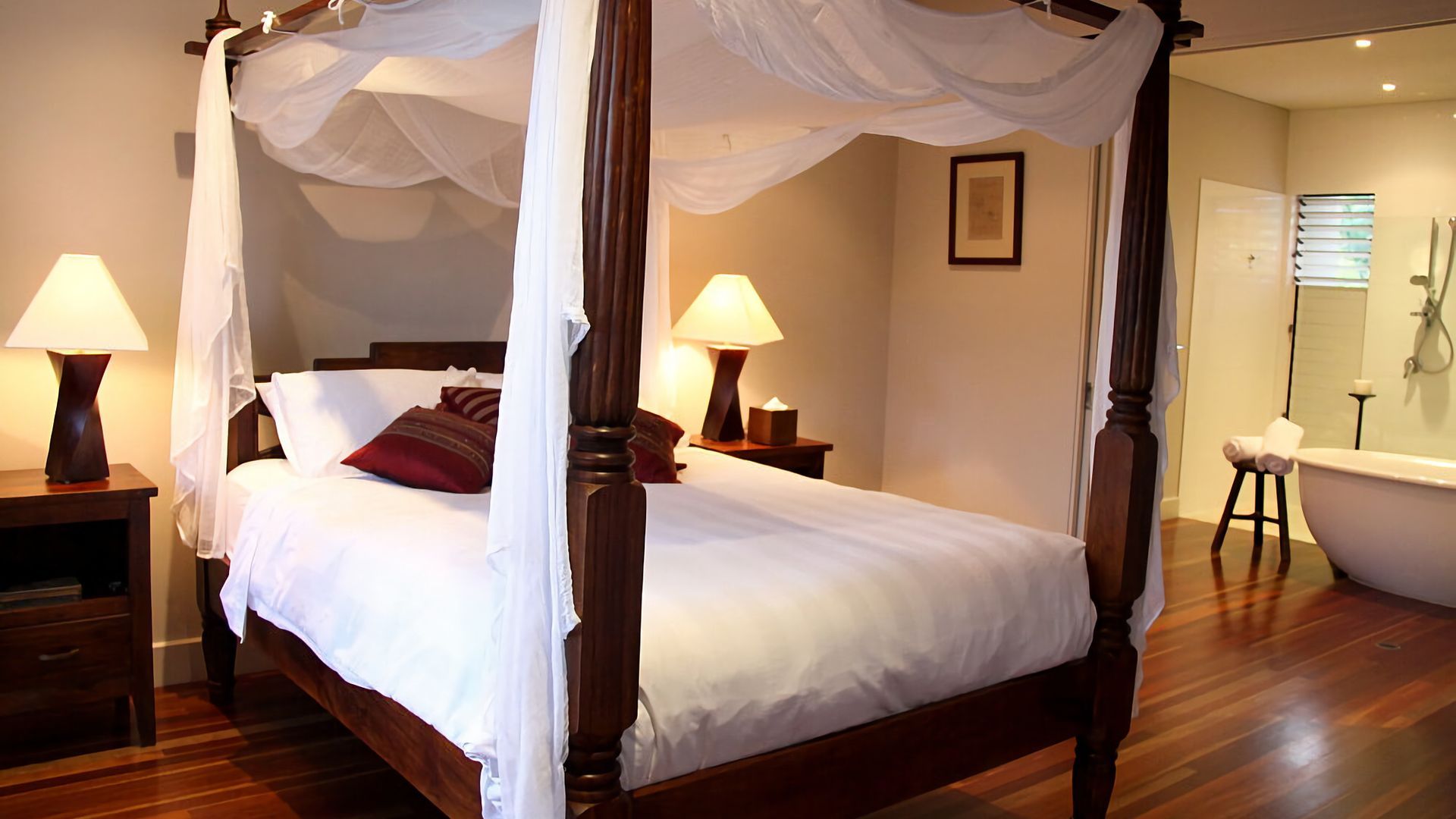 A four-poster canopy bed with white linens and red pillows, two bedside lamps, a wooden nightstand, and a wooden floor.