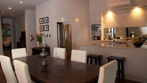 A modern kitchen with stainless steel appliances, a dining area with a wooden table and white chairs, and a living space visible in the background.
