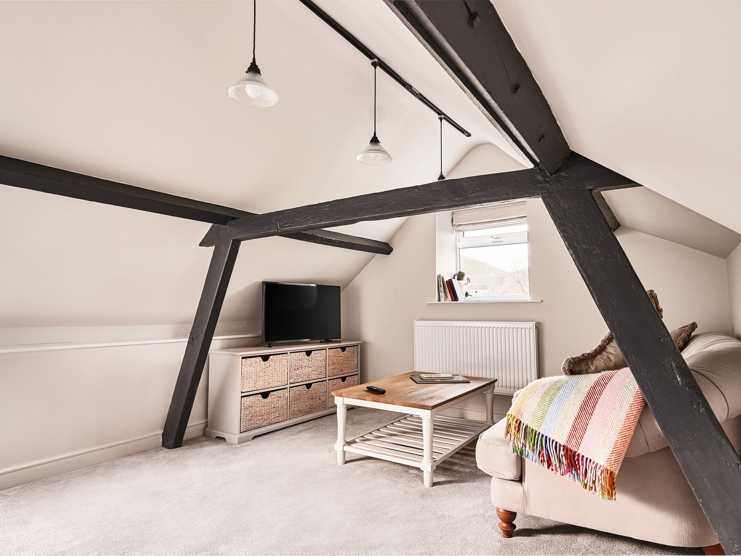 Une pièce mansardée chaleureuse, avec un plafond en pente, un téléviseur posé sur un meuble blanc, une table basse et un canapé agrémenté d’un plaid aux couleurs vives.