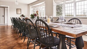 Une salle à manger avec une longue table dressée pour six personnes, entourée de chaises noires, dotée de grandes fenêtres et d’un luminaire suspendu.