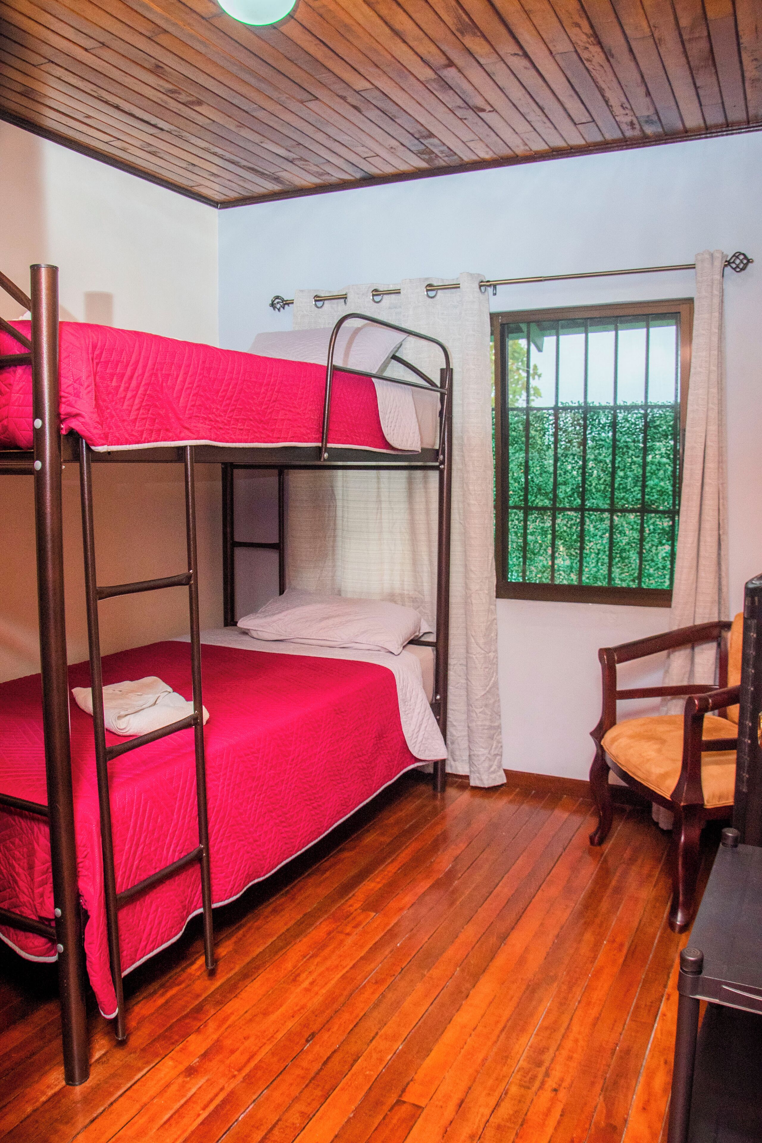 A bunk bed room with a wooden floor, a window with bars, and a chair.