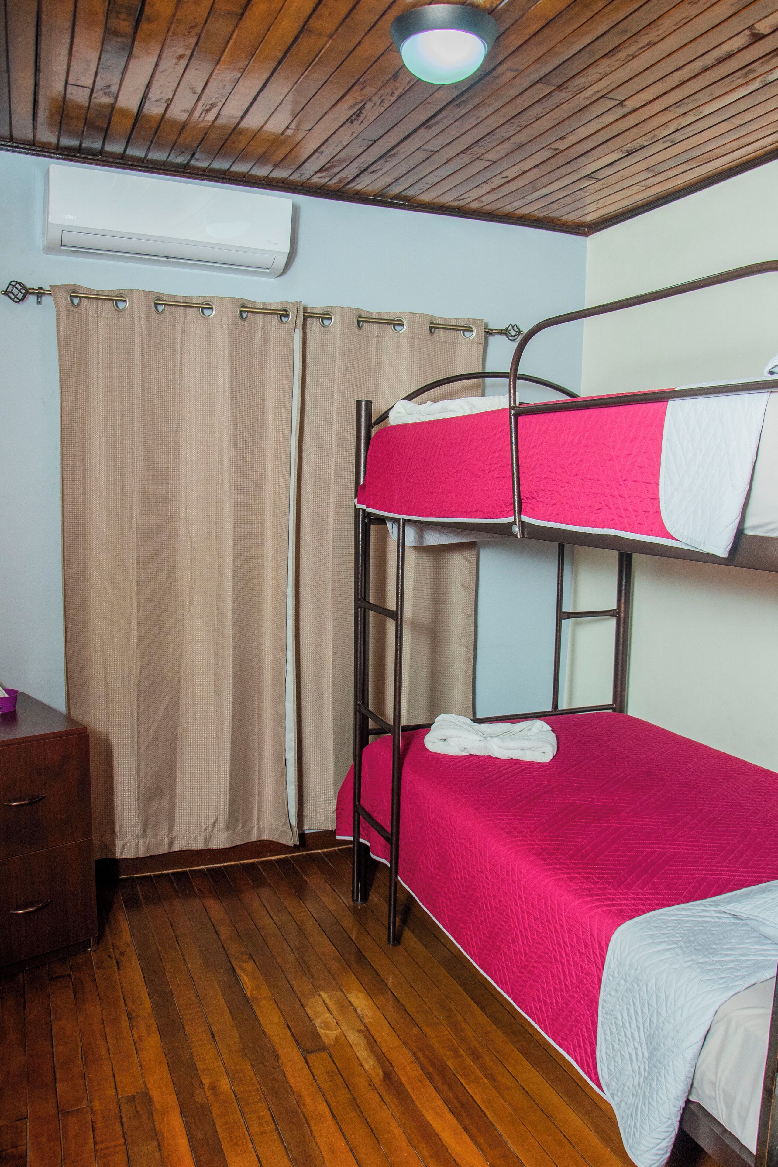 A bunk bed room with a wooden ceiling, an air conditioning unit, a wooden dresser, and a curtain.