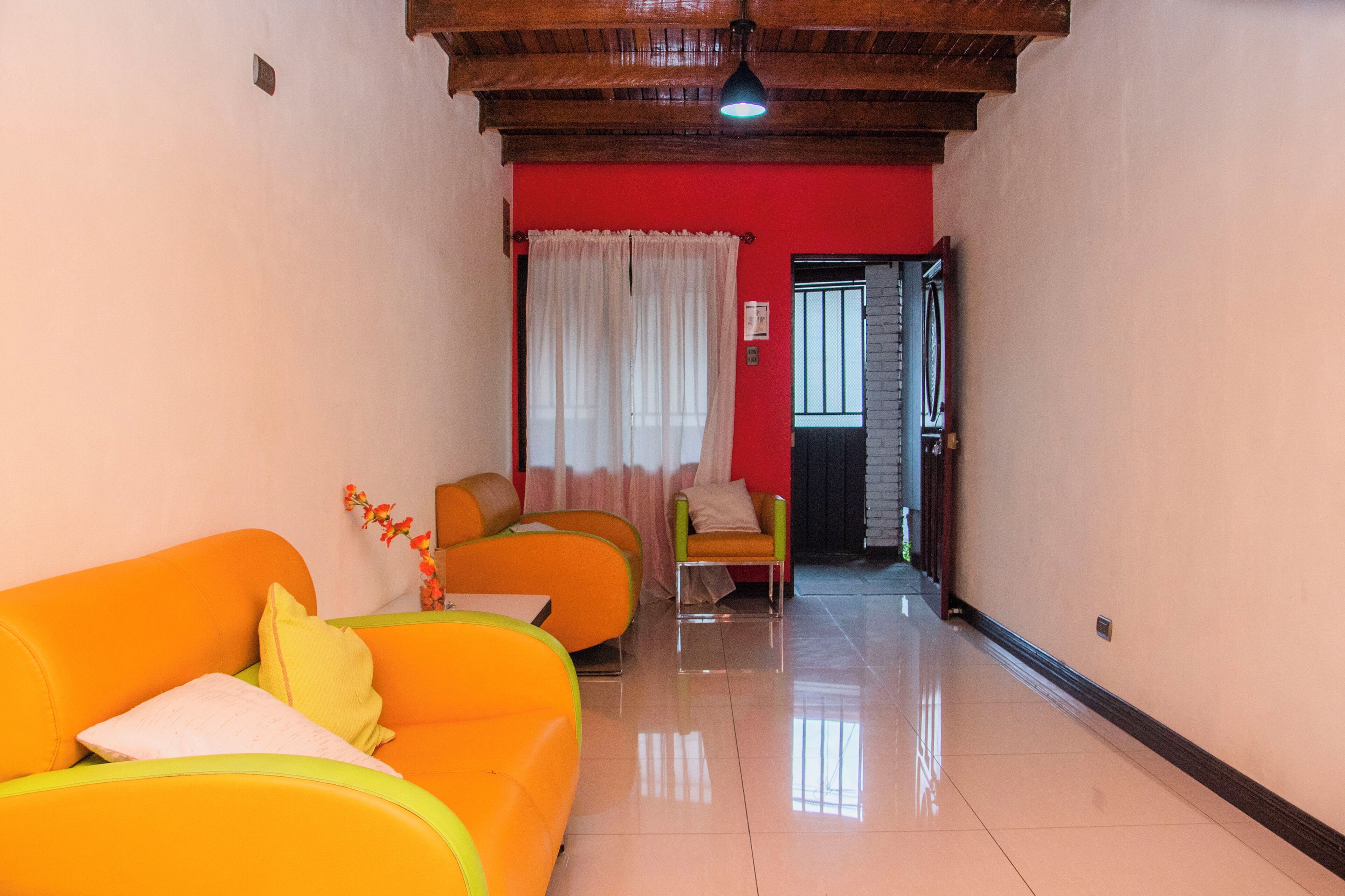 Un pasillo con una pared roja, dos sofás naranjas, un sillón verde y una cortina blanca.