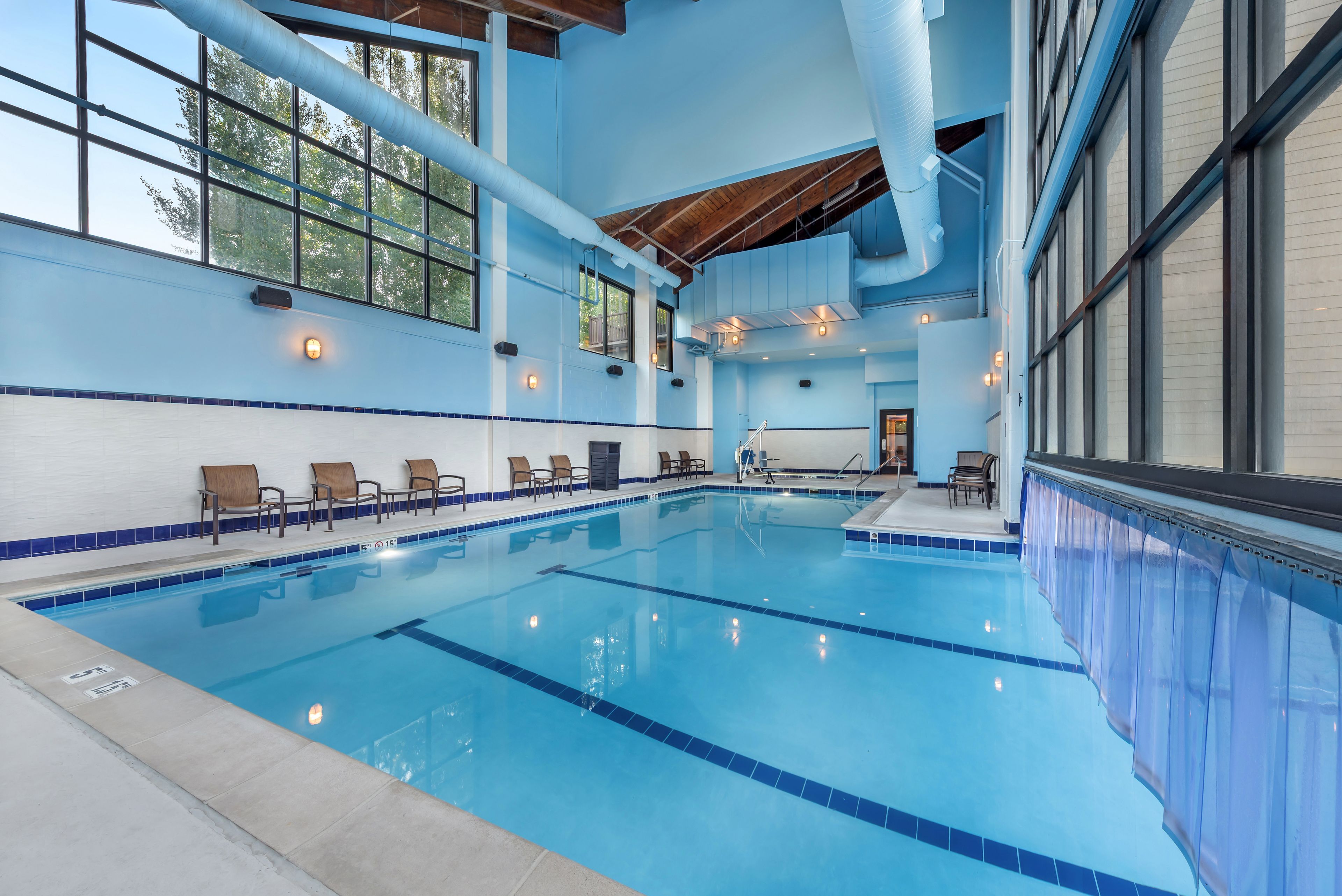 An indoor swimming pool with clear blue water, surrounded by blue-tiled walls and wooden chairs.