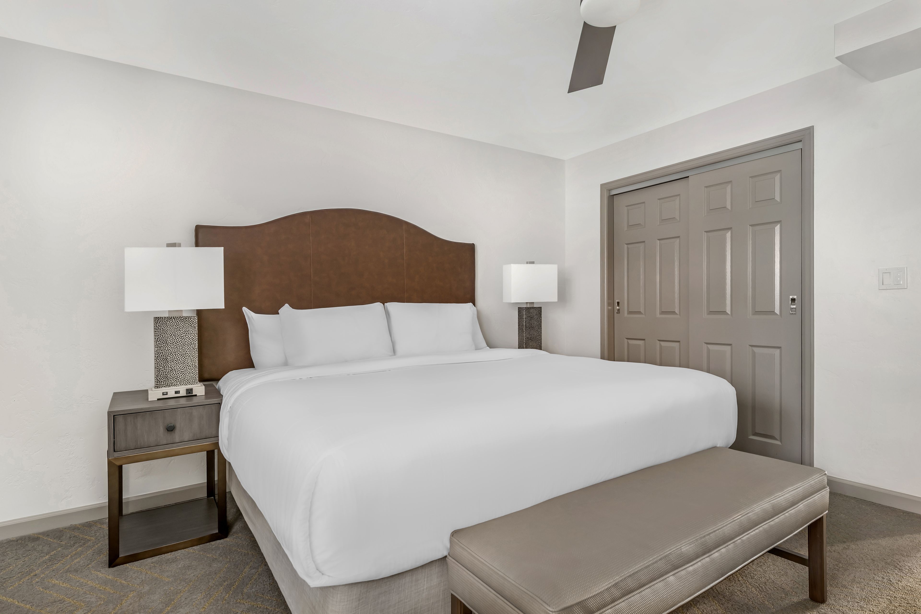 Una habitación de hotel con una cama grande, dos mesitas de noche con lámparas, un banco y una puerta.