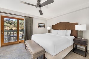 A hotel room with a large bed, a bedside table with a lamp, a bench, a ceiling fan, and a sliding glass door leading to an outdoor area.