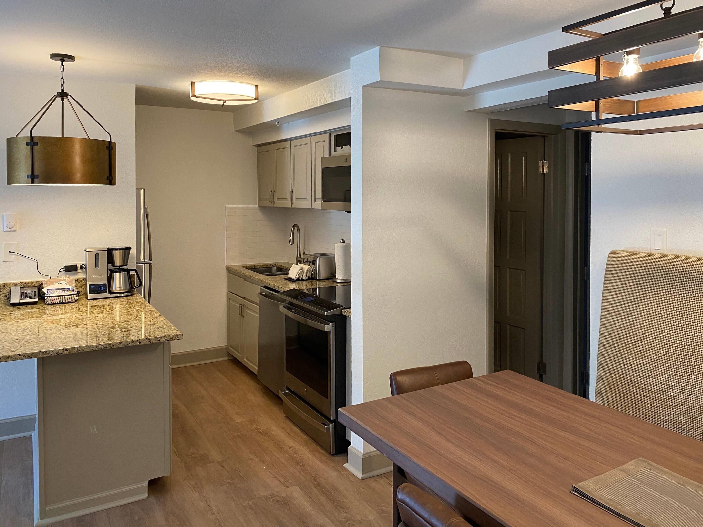 A compact kitchen with a coffee maker, toaster, and oven, a dining table with chairs, and a hanging light fixture.