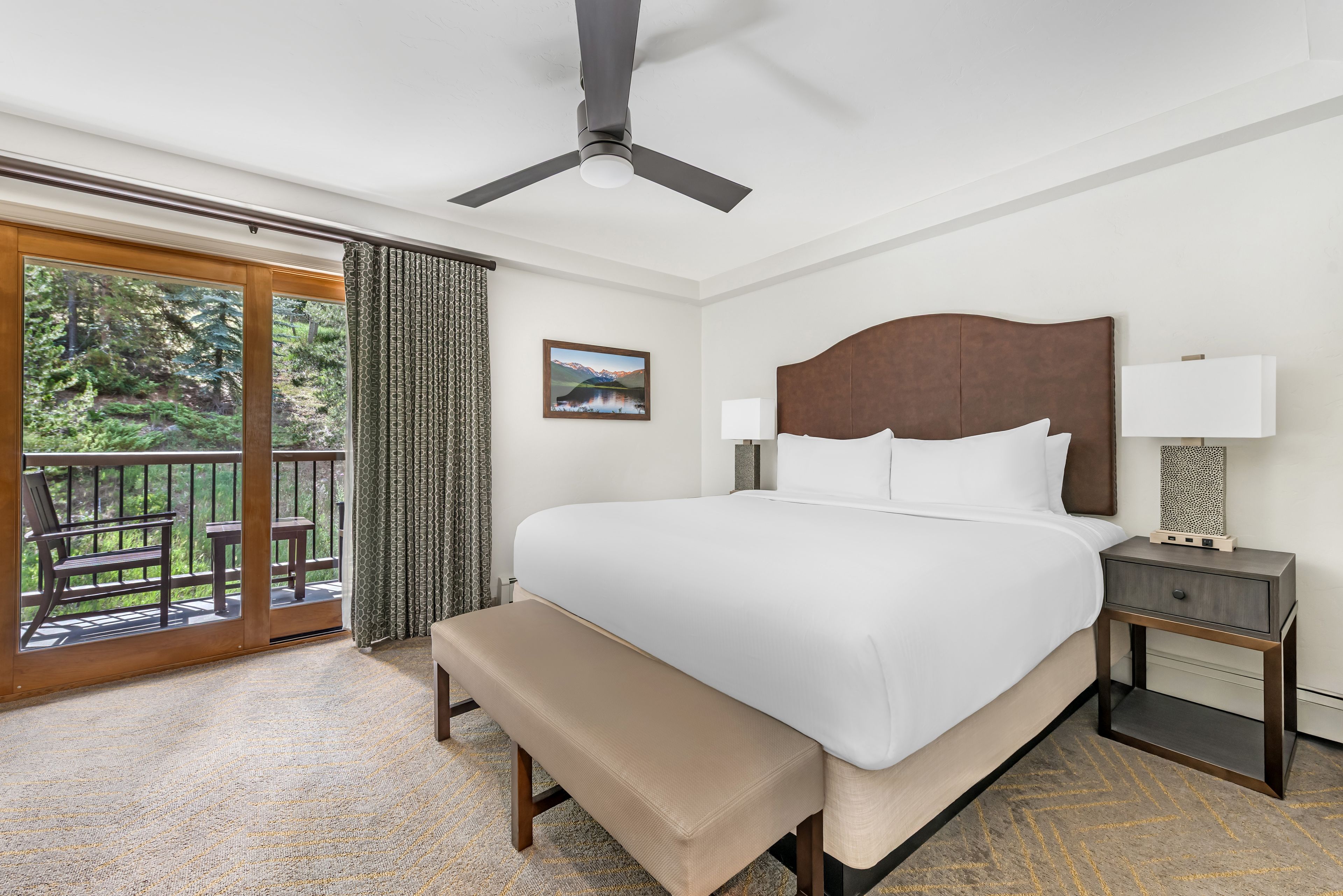 A hotel room with a bed, a bench, a nightstand, a lamp, a picture on the wall, and a balcony with a view of greenery.