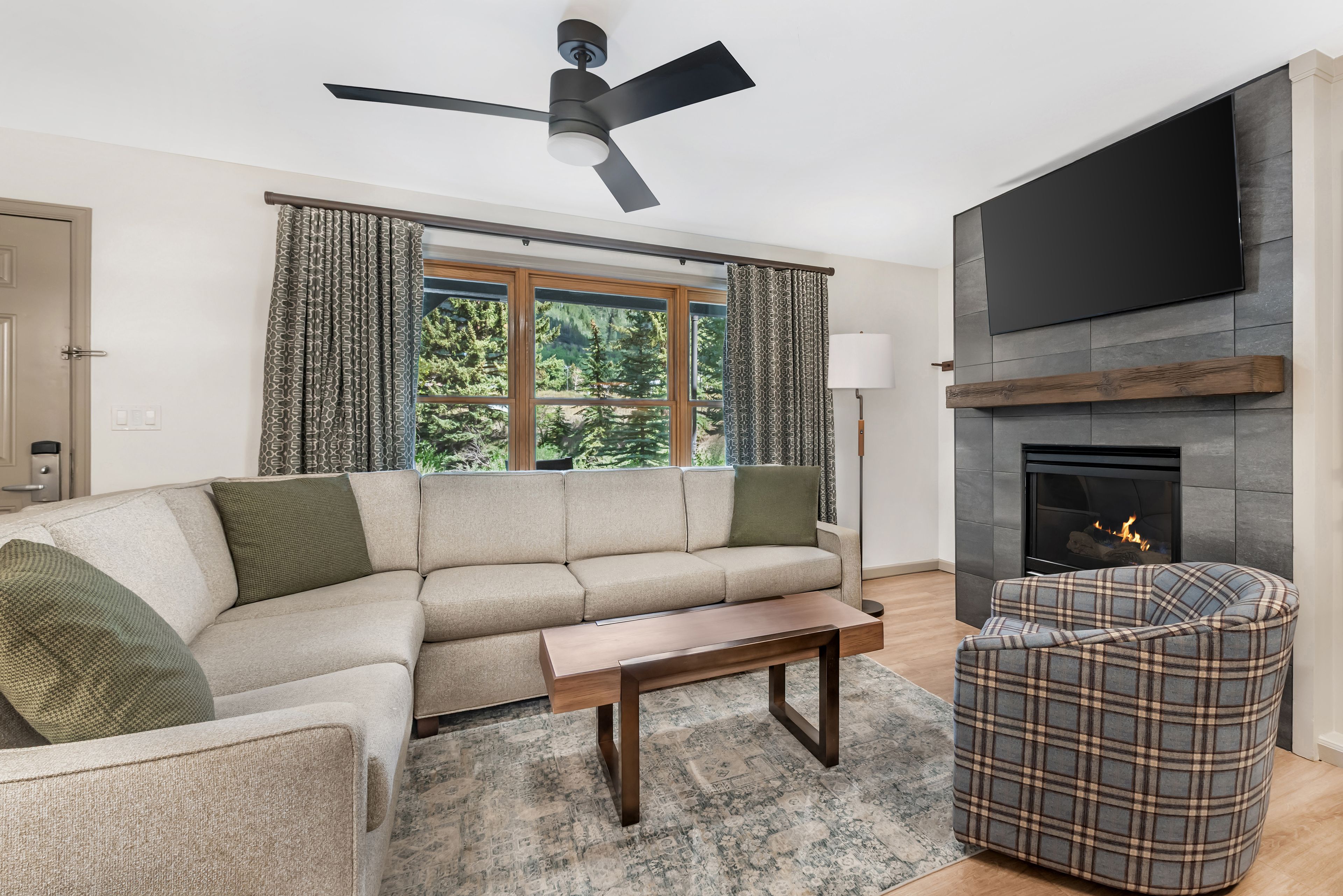 A modern living room with a sectional sofa, a plaid armchair, a fireplace, and a ceiling fan.