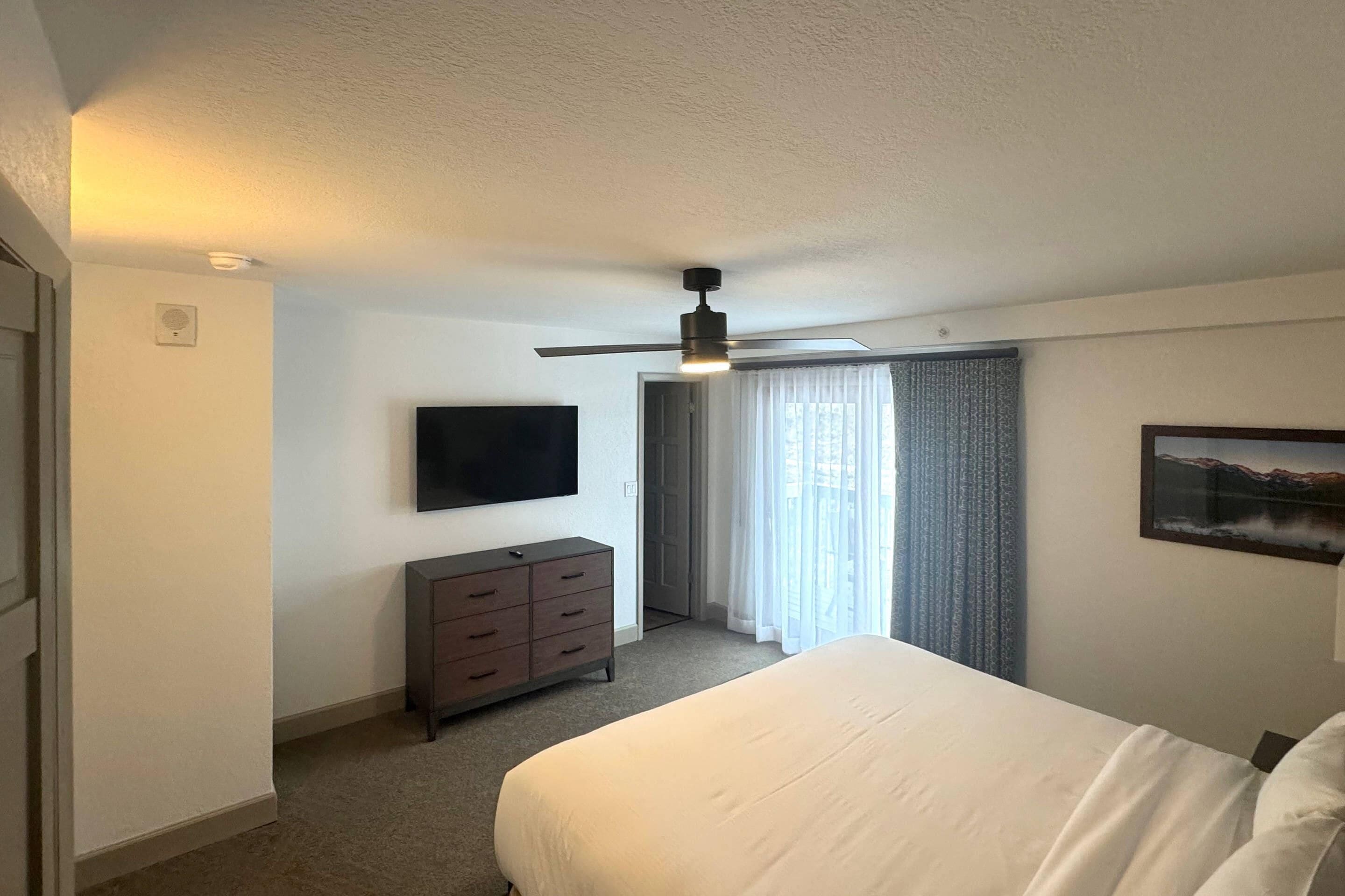 A hotel room with a bed, a TV, a dresser, a ceiling fan, and a door leading to a balcony.