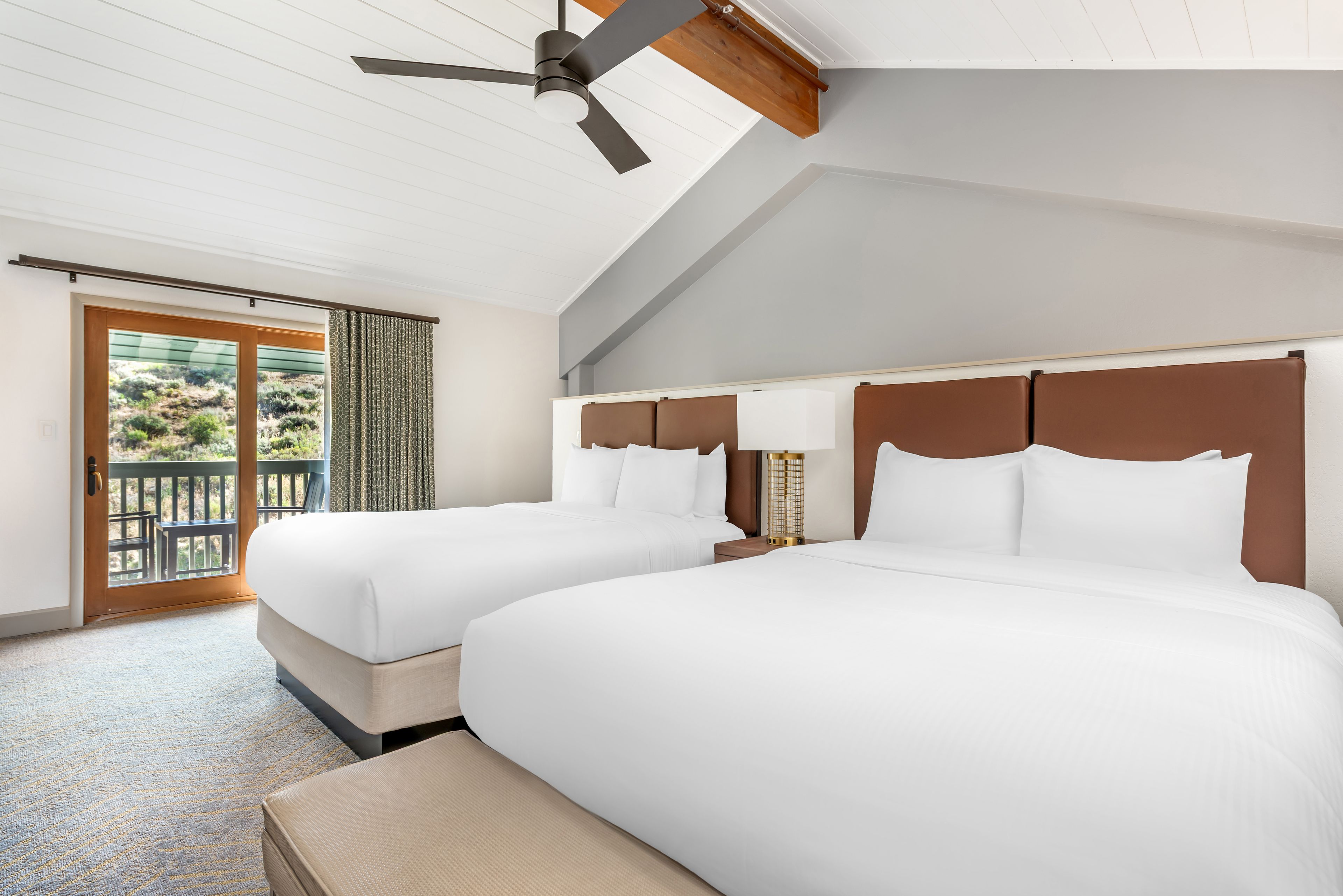 Habitación de hotel con dos camas, ventilador de techo, balcón con vistas y un gran ventanal.