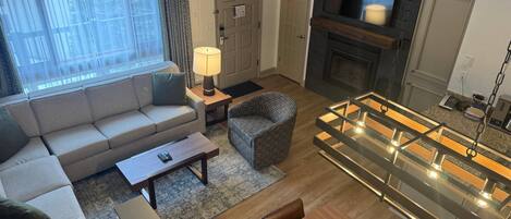 A modern living room with a sofa, a plaid armchair, a glass-top dining table, a fireplace, and a television.