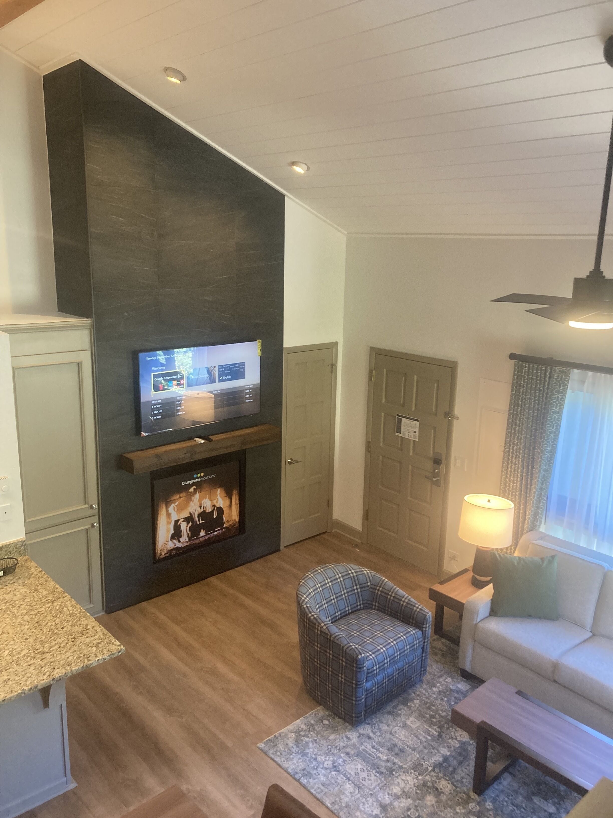 Een woonkamer met een open haard, een televisie erboven, een geruit fauteuil, een bank, een bijzettafeltje met een lamp en een deur.