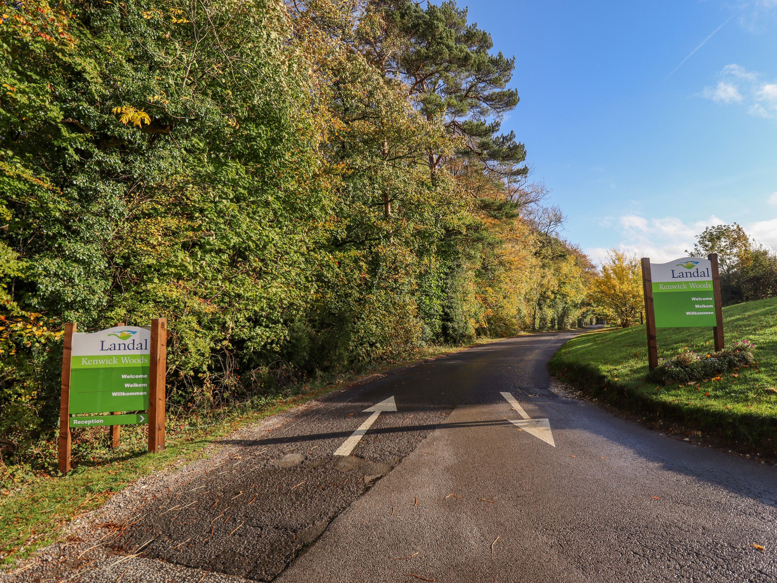Una strada che si inoltra in una zona boschiva, con un cartello che indica 'Landal Kenwick Woods' e 'Benvenuti'.