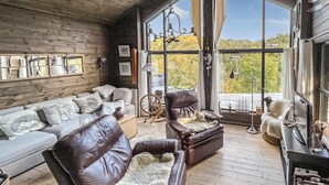 Un interior acogedor con techo de madera, un sofá, un sillón de cuero y un amplio ventanal con vistas a árboles.
