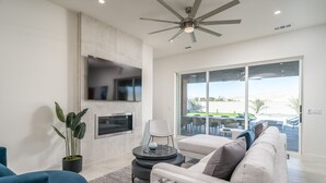 Ein modernes Wohnzimmer mit einem Flachbildfernseher, Deckenventilator, Kamin und einer großen Glasschiebetür, die auf eine Außenpatio führt.