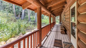 Une cabane en rondins de bois, dotée d’un porche couvert, de grandes fenêtres et offrant une vue sur une zone boisée.