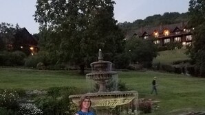 Eine Frau steht vor einer mehrstufigen Fontäne in einem angelegten Garten mit einem großen Baum und einem Gebäude im Hintergrund.