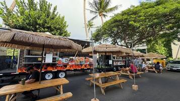 Un camion-restaurant avec un menu et un grill, des tables en bois et des parasols en chaume pour les clients qui souhaitent manger à l’extérieur, et des palmiers en arrière-plan.
